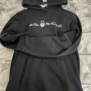 Svart hoodie från Sail Racing - Snygg svart hoodie från Sail Racing med vit logga på bröstet. Den har en klassisk känguruficka och en bekväm huva. Perfekt för en avslappnad stil.  Pris kan diskuteras!!