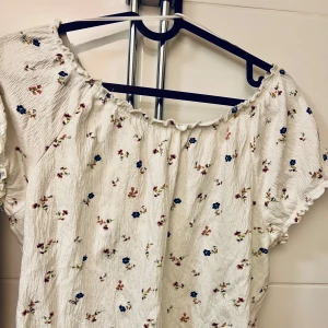 Blommig offshoulder blus - Charmig vit blus med små blommor i blått och rött. Offshoulder-design med korta ärmar och lätt rynkad halslinje. Perfekt för en somrig look!