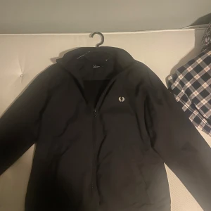 Svart jacka från Fred Perry - Säljer en stilren svart jacka från Fred Perry med dragkedja och diskret logga på bröstet. Jackan har långa ärmar och en klassisk krage. Perfekt för en minimalistisk look. Kan även acceptera bud