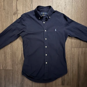 Ralph Lauren skjorta - Säljer nu en snygg mörkblå skjorta från Ralph Lauren i storlek S.🤩 Tillverkad i 100% bomull med stretch för extra komfort. Skjortan har långa ärmar och klassiska knappar framtil. Perfekt för en stilren look nu till våren. Passar både till vardags och lite finare tillfällen.💛Bara hör av dig ifall du har några frågor eller funderingar.🤝