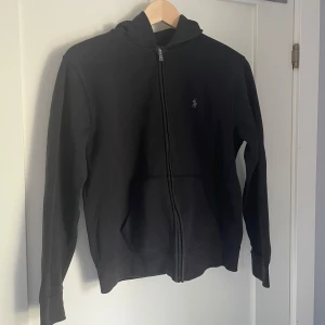 Svart hoodie från Ralph Lauren - Säljer en stilren svart hoodie från Ralph Lauren med dragkedja och broderad logotyp på bröstet. Perfekt för en avslappnad look. Hoodien har långa ärmar och en klassisk passform.