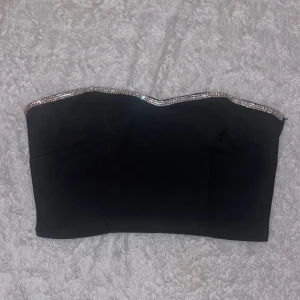 Svart tubtopp från H&M med glittrig kant - Snygg svart tubtopp från H&M med en glittrig kant upptill. Perfekt till fest eller annan tillställning! Den har en elastisk baksida för bättre passform. Använd 1 gång!