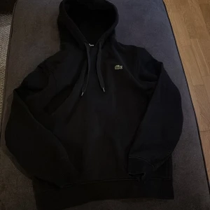 Svart hoodie från Lacoste Sport - Säljer en stilren svart hoodie från Lacoste Sport. Den har en klassisk design med dragsko i huvan och den ikoniska krokodilloggan på bröstet. Perfekt för en avslappnad look.