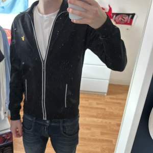 Svart tröja från Lyle & Scott - Snygg svart track jacket från Lyle & Scott med dragkedja. Jackan har en gul broderad logga på bröstet och vita detaljer längs dragkedjan. ”Perfekt” size till 177 CM