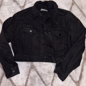Svart croppad jeansjacka - Snygg svart croppad jeansjacka med knappar framtill och två bröstfickor. Perfekt för en cool och avslappnad stil. Jackan har långa ärmar och en klassisk krage.