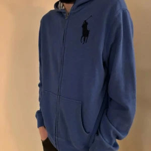 Blå hoodie med dragkedja från Ralph Lauren - Xl barn storlek passar S Säljer en snygg blå hoodie från Ralph Lauren med dragkedja och det klassiska logotypen på bröstet. Tröjan har långa ärmar och en bekväm passform. Perfekt för en avslappnad stil.