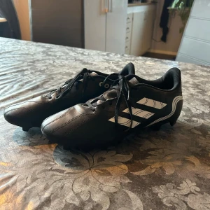 Svarta fotbollsskor Adidas Copa - Säljer ett par svarta Adidas Copa fotbollsskor med vita detaljer och snörning. Skorna har en klassisk design med tre vita ränder på sidan och är perfekta för fotbollsplanen. Dom är använda en gång. 