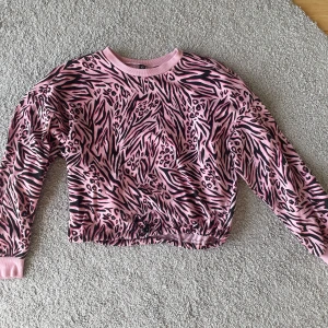 Rosa och svart mönstrad tröja - Säljer en cool rosa och svart mönstrad tröja med djurprint. Tröjan har långa ärmar och en justerbar dragsko i nederkanten för en perfekt passform. Perfekt för att ge din outfit en färgglad touch!