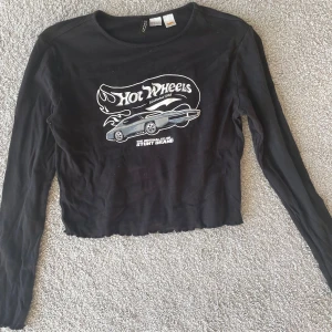 Svart långärmad Hot Wheels tröja - Säljer en cool svart långärmad tröja från Hot Wheels med ett tryck av en bil och logotyp på framsidan. Tröjan är croppad och har en avslappnad passform. Perfekt för dig som gillar en unik stil!