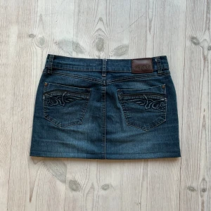 Blå lågmidjad jeanskjol  - Mörkblå jeanskjol från Angels som jag har sytt upp från en längre kjol. Kan inte hitta storlek men skulle säga S/M. Tvärs över midjan är det 38 cm och längs med sidan är det ca 32 cm. Fint skick. Använd gärna köp nu!