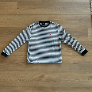 Randig långärmad tröja med logga - Säljer en stilren randig tunnare Axel Arigato sweatshirt  med svart och vit design. Tröjan har en liten röd fågel på bröstet och svarta muddar vid ärmslut och hals. Perfekt för en avslappnad look.