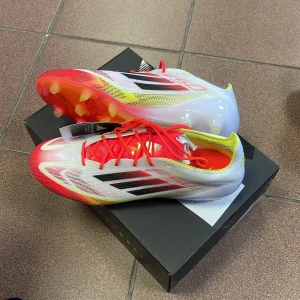 Adidas fotbollsskor med röda detaljer - Helt nya aldrig använda. Väldigt snygga fotbollsskor från Adidas i vitt med röda och gula detaljer. Skorna har en modern design med svarta ränder och snörning. Storlek 45, kvitto finns och lådan och påse