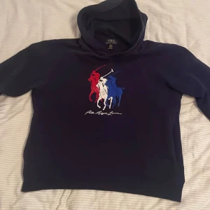 Mörkblå hoodie från Polo Ralph Lauren - Säljer en snygg mörkblå hoodie från Polo Ralph Lauren med ett coolt tryck av tre polospelare i rött, vitt och blått på framsidan. Hoodien är tillverkad i en mjuk bomullsblandning och har en klassisk passform med långa ärmar och en bekväm huva. Den är i storlek xl i barn storlek men sitter som en S köpt på zalando har ej kvitto pris kan diskuteras 