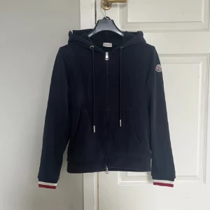 Mörkblå hoodie från Moncler - Snygg svart hoodie från Moncler med dragkedja och justerbar huva. Tröjan har Moncler-loggan på ärmen och ribbade muddar med röd och vit detalj vid ärmslut. Perfekt för en stilren och bekväm look. Tröjan är storlek s men passar XS! Köpte denna på Plick så vet ej om den är äkta kan tänka mig gå ner lite i pris vid snabb affär