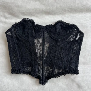 Svart spetskorsett - Snygg svart korsett i spets med hakknäppning bak. Den har en elegant design med spetsdetaljer och ger en smickrande passform. Perfekt för att lägga till en touch av stil till din outfit.