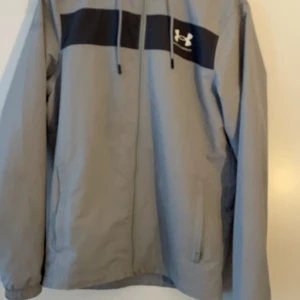 Grå vindjacka från Under Armour - Säljer en grå vindjacka från Under Armour med svart detalj över bröstet och logotyp. Jackan har dragkedja och långa ärmar, perfekt för blåsiga dagar. Den är stilren och funktionell med en sportig look. Defekterna syns på bilden