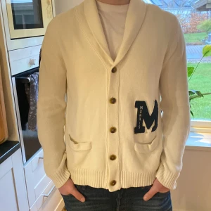 Vit cardigan från Morris - Säljer en stilren vit cardigan från Morris med knappar framtill och två fickor. Koftan har en stor M-logga på ena sidan och en etikett med brittisk flagga på ärmen. Perfekt för en avslappnad och snygg look.