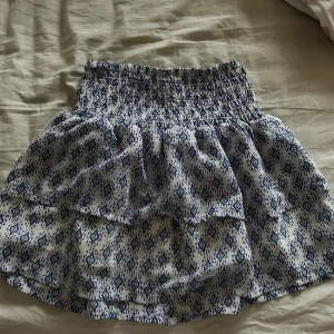 Blåmönstrad kortkjol med volang - Charmig kortkjol med blått mönster och volangdetaljer. Den har en elastisk midja för bekväm passform. Perfekt för varma dagar och ger en luftig känsla.