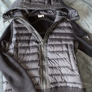 Svart quiltad jacka från Moncler - Snygg svart quiltad jacka från Moncler med huva och dragkedja. Jackan har stickade ärmar och ribbade detaljer. Perfekt för kyligare dagar med sin stilrena design och praktiska fickor.