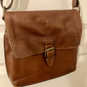 Brun axelväska i skinn - Snygg brun axelväska i skinn med ett klassiskt spänne framtill. Väskan har en praktisk innerficka med dragkedja för säker förvaring. Storlek 30x30