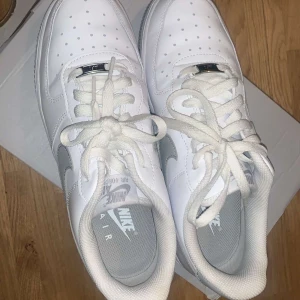 Vita Nike Air Force sneakers - Snygga vita Nike Air Force sneakers med klassisk design och perforerade detaljer på tån. Skorna har en vit sula och grå swoosh-logga på sidan. Perfekta för en stilren look.