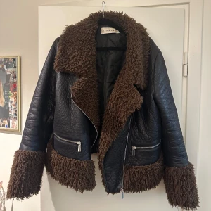 Jacka - Snygg svart skinnjacka från Ivyrevel med mjuka bruna pälsdetaljer på krage, ärmslut och nederkant. Jackan har en cool asymmetrisk dragkedja och två praktiska fickor med dragkedjor. Perfekt för att hålla stilen i kyligare väder.