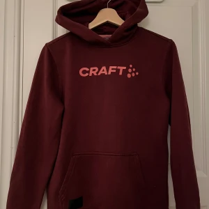 Vinröd hoodie från Craft - Säljer en vinröd hoodie från Craft med en stor ficka framtill och logga i rosa på bröstet. Perfekt för kyliga dagar med sin mysiga huva och långa ärmar. Passar både för avslappnade dagar och träning.