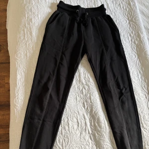 Svarta mjukisbyxor från Lounge Apparel - Snygga svarta mjukisbyxor från Lounge Apparel med elastisk midja och snörning. Byxorna har en avslappnad passform och texten 'LOUNGE' längs benet. Perfekta för en bekväm dag hemma eller en avslappnad utflykt.