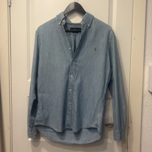 Ljusblå skjorta från Ralph Lauren - Snygg ljusblå skjorta från Ralph Lauren i slim fit. Skjortan har långa ärmar och är tillverkad i ett mjukt bomullsmaterial. Den har knappar framtill och en broderad logotyp på bröstet. Perfekt för en stilren look!