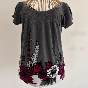 Grå blommig topp från H&M Stl s - Säljer en grå topp från H&M med ett vackert blommönster. Toppen har korta ärmar och en smockad detalj vid halsringningen. Perfekt för en färgglad touch i garderoben!