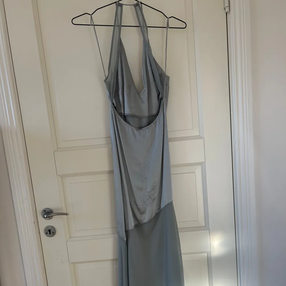 Elegant ljusblå långklänning med djup v-ringning och tunna axelband. Klänningen har en stilren design med en mjuk drapering framtill och en öppen rygg. Perfekt för en somrig kväll eller speciella tillfällen.. Mekot.