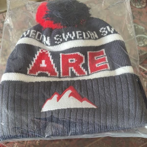 Mössa med texten Åre - Snygg mössa i mörkblått med vit och röd text som säger 'Åre'. Den har en stor tofs i rött och blått på toppen och ett bergmotiv framtill. Perfekt för kalla dagar med en chill outfit, priset ej hugget i sten!