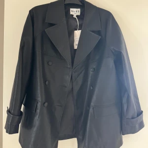 Trenchcoat i skinn imitation - NA-KD - Svart trenchcoat i skinn imitation från NA-KD. Ny med prislapp kvar. Endast testad. Den är oversized.