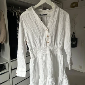 Vit klänning med knappar - Säljer en stilren vit klänning med v-ringning och knappar framtill. Klänningen har långa ärmar och en smockdetalj i midjan för en fin passform. Perfekt för vår och sommar! Nypris 399kr, prislapp kvar