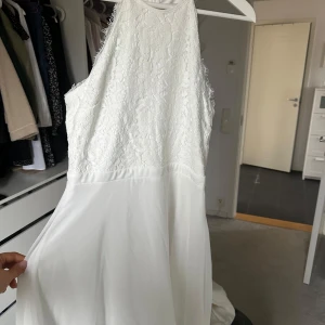 Vit ärmlös klänning med spetsdetaljer - Elegant vit klänning med spetsdetaljer upptill och en flytande kjol. Den är ärmlös och har en hög halsringning, perfekt för sommarens festligheter.