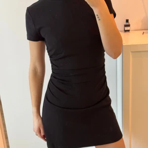 Svart kortklänning från Whitefox  - Snygg svart kortklänning med korta ärmar och en smickrande rynkad detalj i midjan. Tillverkad i ett mjukt och bekvämt material. Dragkedja o rygg, 