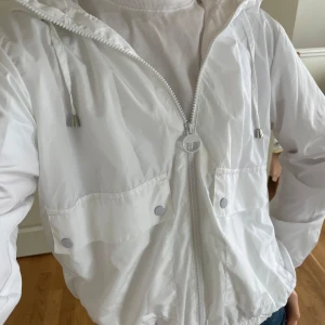 Vit vindjacka med dragkedja - Säljer en stilren vit vindjacka med dragkedja och huva. Jackan har två framfickor med knappar och en elastisk nederkant för en bekväm passform. Perfekt för vår och sommar!