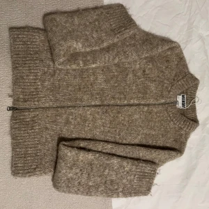 Beige stickad kofta från weekday - Mysig beige stickad kofta från H&M med dragkedja framtill. Perfekt för kyliga dagar med sin varma och bekväma design. Koftan har långa ärmar och ribbade muddar vid ärmslut och nederkant.