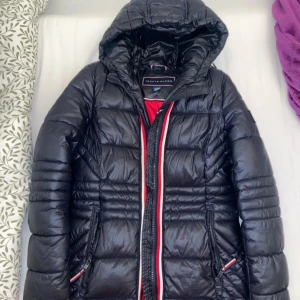  dunjacka från Tommy Hilfiger - Snygg  dunjacka från Tommy Hilfiger med huva och dragkedja. Jackan har röda och vita detaljer längs dragkedjan och på ärmen. Perfekt för kyliga dagar med sin vadderade design.