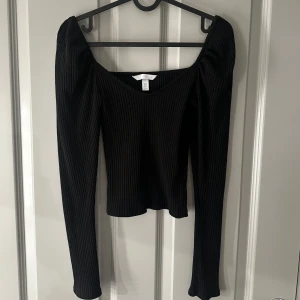 Svart ribbad topp från H&M - Snygg svart ribbad topp från H&M. Den har långa ärmar som är lite puffiga vid axlarna och en stilren design som passar perfekt till olika tillfällen. Toppen är både bekväm och trendig.