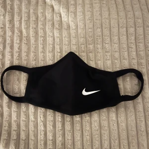 Svart munskydd från Nike - Snygg och stilren svart munskydd från Nike med deras ikoniska logga i vitt. Perfekt för att hålla sig skyddad och samtidigt se sportig ut. Masken har elastiska band för en bekväm passform.