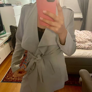 Ljusgrå kappa med knytband - Snygg ljusgrå trenchcoat med knytband i midjan och stora slag. Jackan har långa ärmar och en knappdetalj på baksidan. 