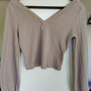 Brun-beige ribbad tröja - Säljer en snygg brun-beige tröja med ribbad struktur och v-ringad design. Tröjan är croppad och har långa ärmar, perfekt för en stilren look. Passar bra till både jeans och kjol.