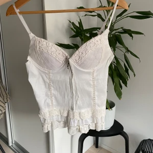 Vit spetsdetaljerad korsettopp - Säljer en elegant vit korsettopp med spetsdetaljer och justerbara axelband. Toppen har en dragkedja framtill och vacker spetskant nedtill. Perfekt för en stilren look.