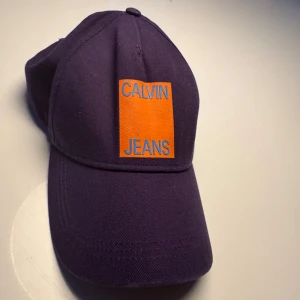 Lila keps från Calvin Klein Jeans - Snygg lila keps från Calvin Klein Jeans med en iögonfallande orange patch och blå text framtill. Kepsen har en klassisk design med böjd skärm och justerbar passform.
