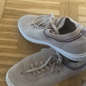 Beige sneakers från Tommy Hilfiger - Snygga beige sneakers från Tommy Hilfiger med snörning och diskret logga på sidan. Skorna har en ribbad kant och en bekväm sula med märkets namn på undersidan. Perfekta för en stilren vardagslook.