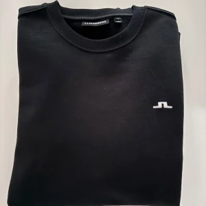 Svart tröja från J.Lindeberg - Snygg svart tröja från J.Lindeberg med rund hals och diskret logga på bröstet. Perfekt för en stilren look. Tröjan har långa ärmar och ribbade muddar vid ärmslut och nederkant. Bara att skriva om man har några funderingar!