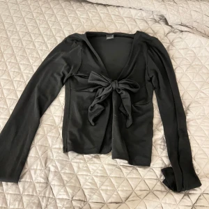 Svart långärmad topp från Gina Tricot - Snygg svart långärmad topp från Gina Tricot med en stor knytrosett framtill. Perfekt för en stilren look. Toppen har en djup v-ringning och är tillverkad i ett mjukt material.