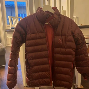 Röd dunjacka från Haglöfs - Säljer en röd dunjacka (kan använda båda sidorna) från Haglöfs med dragkedja framtill. Jackan har en quiltad design och långa ärmar, perfekt för kyligare väder. Den är stilren och funktionell, idealisk för både stad och natur.