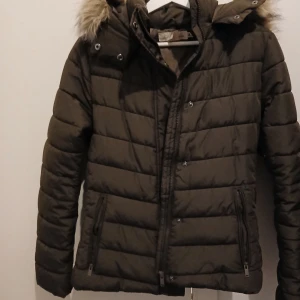 Brun dunjacka från H&M - Säljer en brun dunjacka från H&M med pälskantad huva. Jackan har dragkedja och knappar framtill samt två fickor med dragkedja. Perfekt för kyliga dagar!